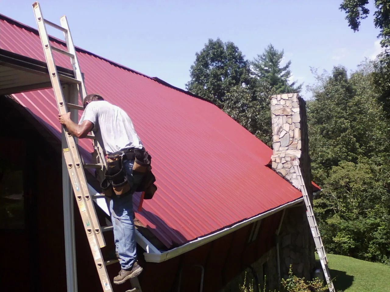 Roofer on a ladder beside a red metal roof and stone chimney — Soffit & Fascia Repair by Roofing Brothers in Dundalk